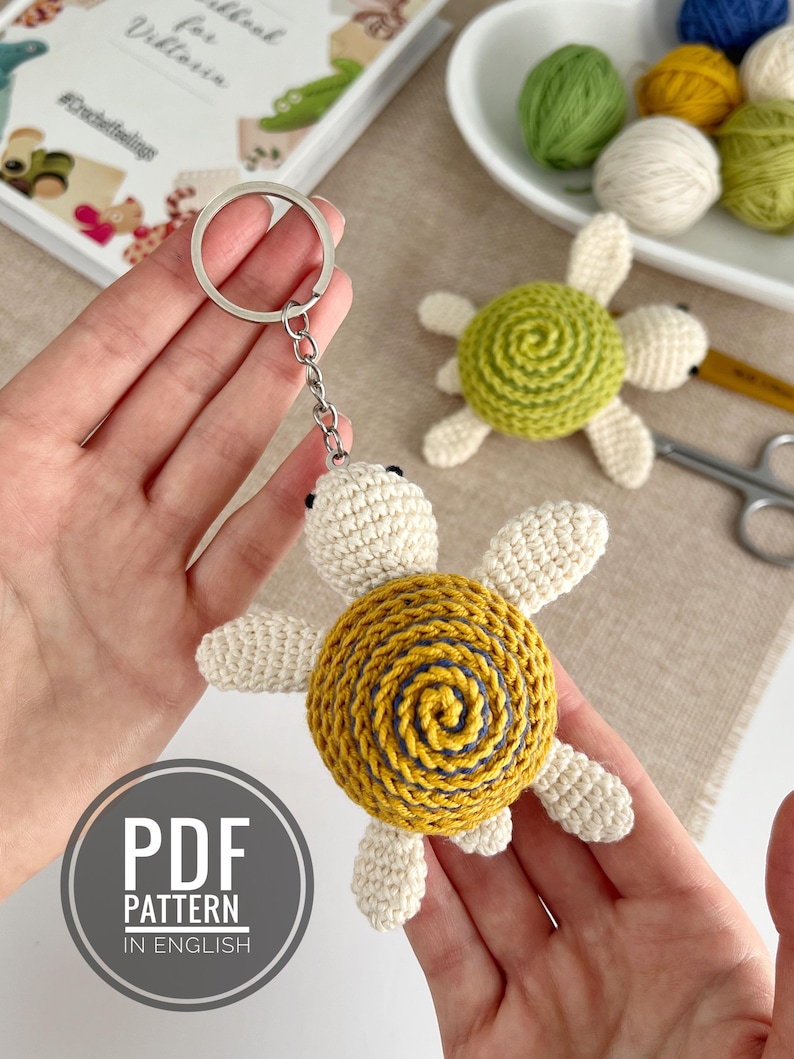 Puede incluir: Llavero de tortuga de ganchillo con caparaz&oacute;n amarillo y azul, cabeza y extremidades color crema y un llavero plateado. La imagen tambi&eacute;n muestra un patr&oacute;n PDF en ingl&eacute;s y otras tortugas de ganchillo.