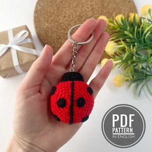 May include: A red and black crocheted ladybug keychain. The keychain features a round, red body with black spots and a black head. The keychain is attached to a silver metal ring. The image also includes a gift box and the text "PDF PATTERN IN ENGLISH".
