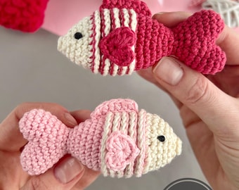 May include: Two handmade crochet fish in shades of pink and red, with white accents and heart-shaped details. The fish have striped patterns and are held in hands. The image includes a "PDF PATTERN IN ENGLISH" logo.