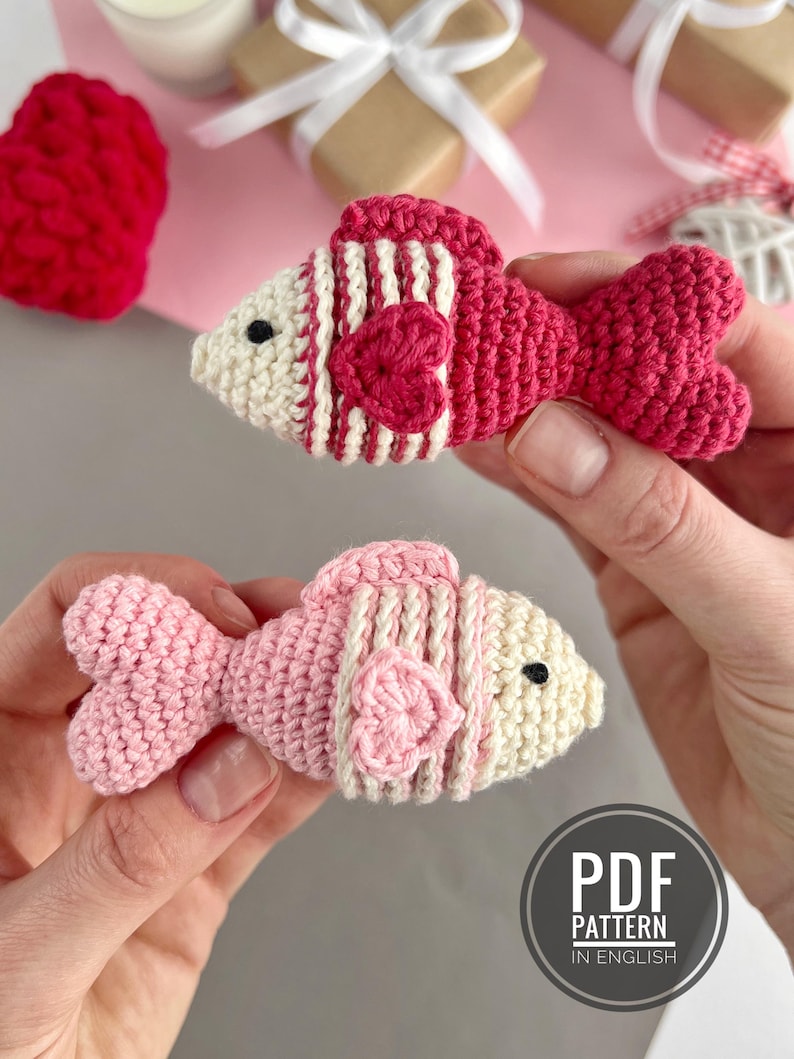 May include: Two handmade crochet fish in shades of pink and red, with white accents and heart-shaped details. The fish have striped patterns and are held in hands. The image includes a "PDF PATTERN IN ENGLISH" logo.