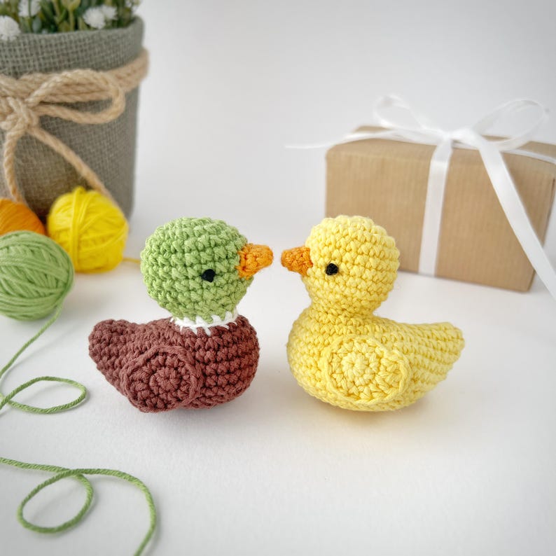May include: Two handmade crochet ducklings. One duckling is yellow with an orange beak and black eyes. The other duckling has a green head, brown body, and orange beak. They are placed on a white surface.