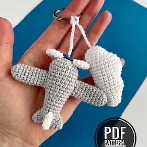 Puede incluir: Llavero de avión tejido a ganchillo en gris con detalles blancos, incluyendo la hélice y la cola. Un charm en forma de nube blanca está adjunto. El llavero es sostenido por una mano sobre un fondo azul. El texto "PDF PATTERN IN ENGLISH" es visible.