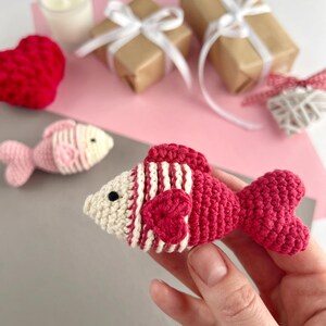 May include: A handmade crochet fish in shades of red and cream, held in a hand. The fish features striped detailing and a heart-shaped accent. Other items in the background include wrapped gifts, a candle, and a crochet heart.