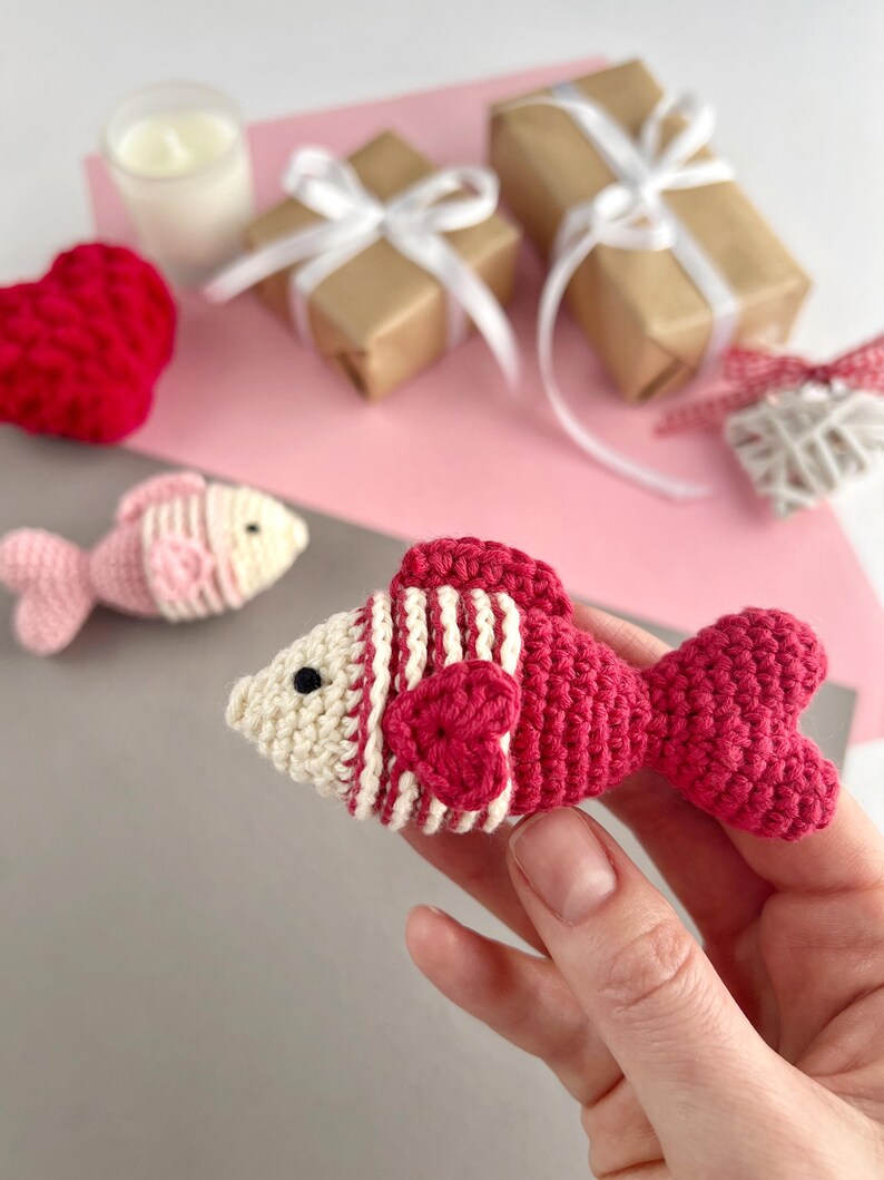 May include: A handmade crochet fish in shades of red and cream, held in a hand. The fish features striped detailing and a heart-shaped accent. Other items in the background include wrapped gifts, a candle, and a crochet heart.
