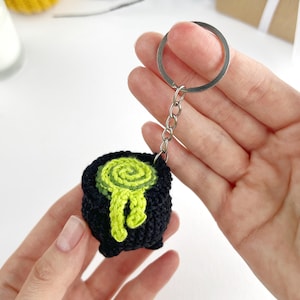 May include: A black crocheted cauldron keychain featuring a bright lime green spiral design on top with a drip effect extending down the side. The small, round, three-legged pot is attached to a silver-toned metal split ring by a short chain.