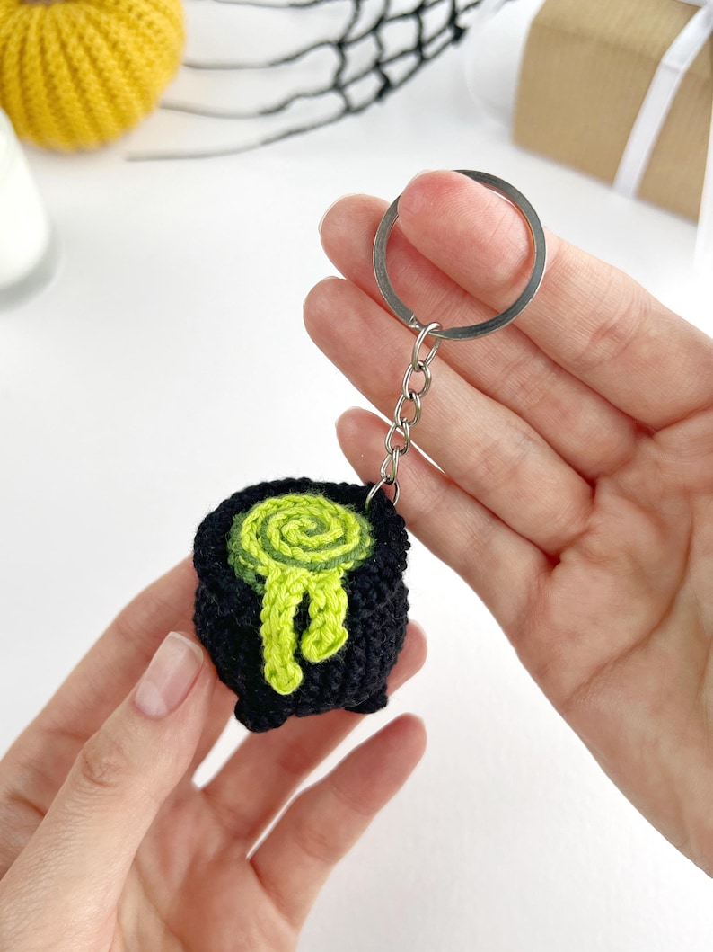 May include: A black crocheted cauldron keychain featuring a bright lime green spiral design on top with a drip effect extending down the side. The small, round, three-legged pot is attached to a silver-toned metal split ring by a short chain.