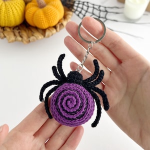 May include: A crocheted spider keychain featuring a round purple body with a black spiral pattern and eight black legs. The spider is attached to a silver-toned metal split ring by a short chain, creating a textured, handmade accessory.
