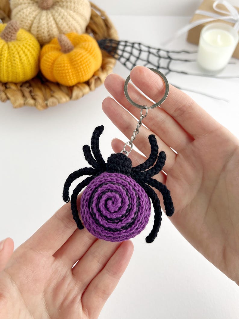 May include: A crocheted spider keychain featuring a round purple body with a black spiral pattern and eight black legs. The spider is attached to a silver-toned metal split ring by a short chain, creating a textured, handmade accessory.