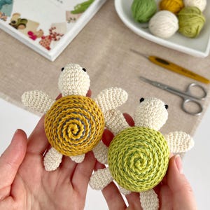 Puede incluir: Dos juguetes de tortuga hechos a mano con ganchillo. Una tortuga tiene un caparaz&oacute;n amarillo con una espiral azul, la otra tiene un caparaz&oacute;n verde. Ambas tortugas tienen cuerpos y aletas de color crema. Un libro titulado "Workbook for Viktoria" est&aacute; en el fondo.