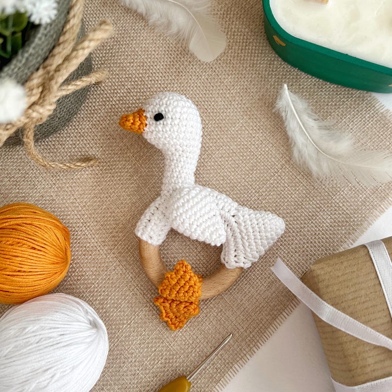 May include: White crocheted goose rattle with a wooden ring and orange crocheted wings. The rattle is on a brown burlap surface.