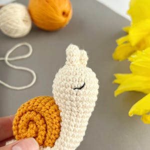 Pode incluir: Um brinquedo de caracol de croch&ecirc; com um corpo branco e uma concha marrom. O caracol tem olhos pretos e a boca fechada.