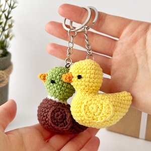 May include: Two handmade crochet duck keychains. One duck is yellow with an orange beak, and the other is green with a brown body. Both keychains have silver metal rings for attaching to keys or bags. The ducks are held in a person's hands.