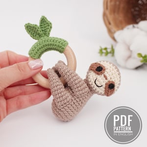 Puede incluir: Sonajero de bebé de crochet con forma de perezoso con un anillo de madera y hojas verdes. El perezoso es marrón y tiene una cara sonriente con ojos marrones. El sonajero es un patrón PDF en inglés.