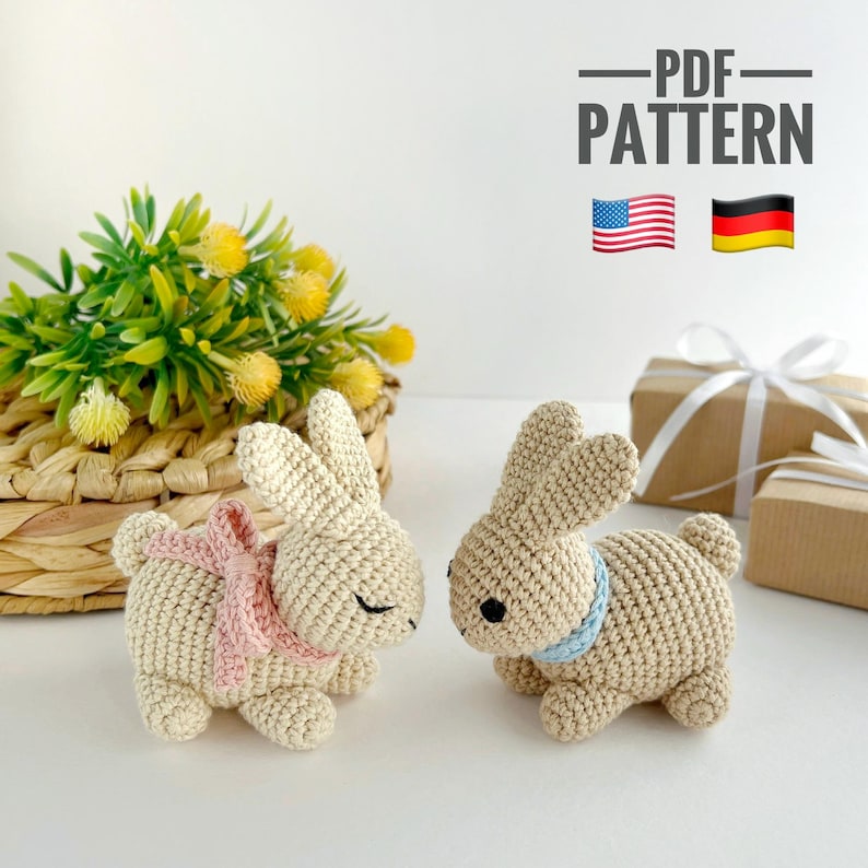 May include: Two crocheted bunny amigurumi toys with a pink and blue ribbon around their necks. The bunnies are sitting on a white surface with a wicker basket and green plants in the background. The image includes the text "PDF PATTERN" and flags for the United States and Germany.