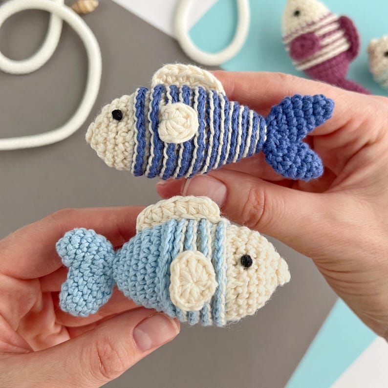 May include: Two handmade crochet fish toys. One fish is blue and white with a blue tail. The other fish is light blue and white with a light blue tail. Both fish have a cream-colored belly and fins. The fish are held in a person's hands.