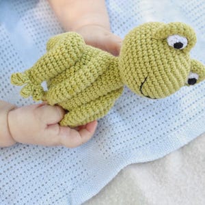 Op de afbeelding: Een handgemaakt, lichtgroen gehaakt kikker speelgoed met witte en zwarte ogen. De kikker ligt op een lichtblauwe deken. Het speelgoed is ontworpen voor baby's en jonge kinderen.