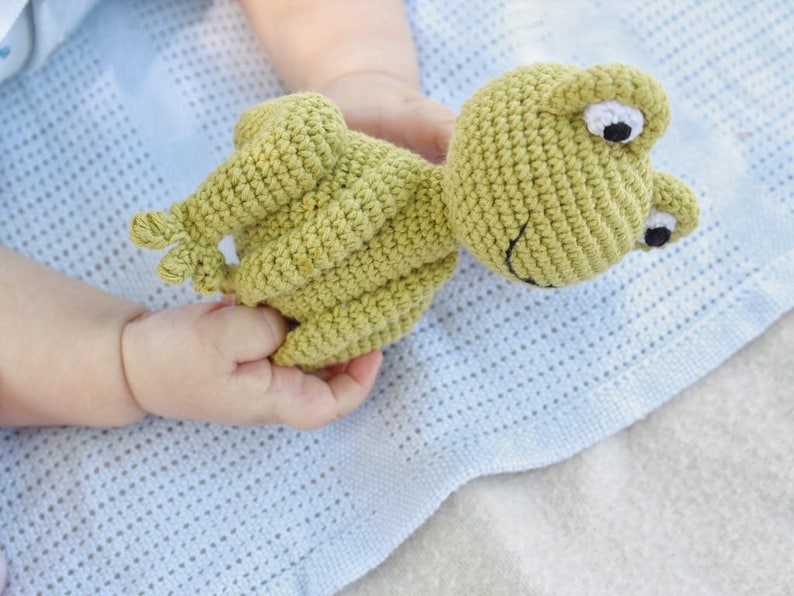 Op de afbeelding: Een handgemaakt, lichtgroen gehaakt kikker speelgoed met witte en zwarte ogen. De kikker ligt op een lichtblauwe deken. Het speelgoed is ontworpen voor baby's en jonge kinderen.
