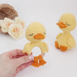 Puede incluir: Dos juguetes de pato amarillo de crochet. Un pato es un sonajero con un anillo de madera y patas naranjas. El otro pato est&aacute; sentado sobre una superficie blanca.