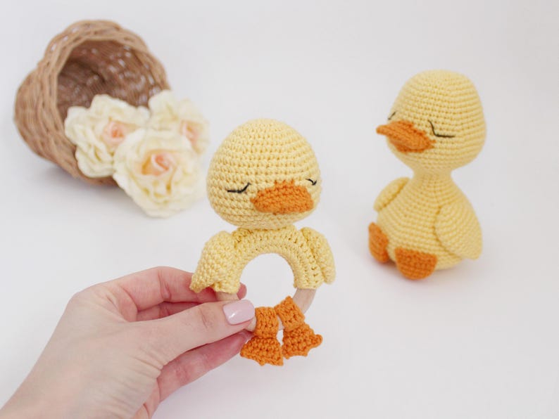 Puede incluir: Dos juguetes de pato amarillo de crochet. Un pato es un sonajero con un anillo de madera y patas naranjas. El otro pato est&aacute; sentado sobre una superficie blanca.
