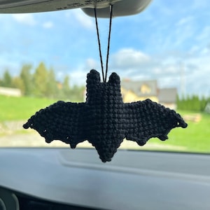 May include: A black, crocheted bat car charm hanging from a black string. The bat has outstretched wings and a textured surface. The background shows a car interior, a blue sky, and trees.