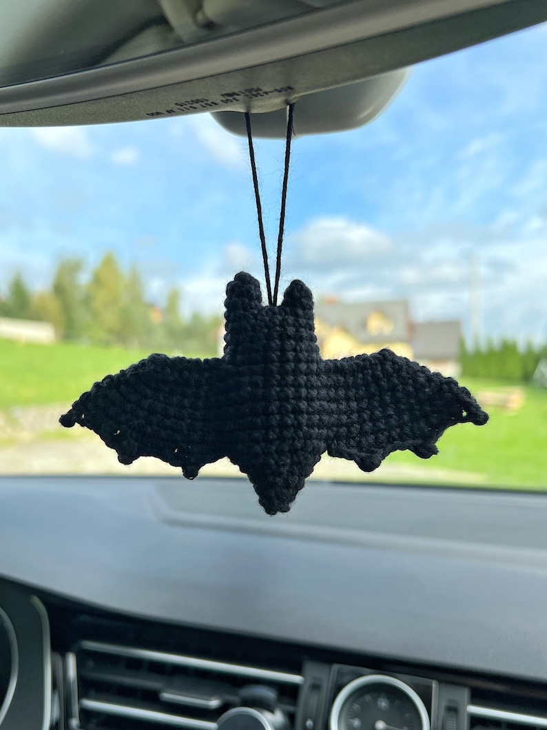 May include: A black, crocheted bat car charm hanging from a black string. The bat has outstretched wings and a textured surface. The background shows a car interior, a blue sky, and trees.