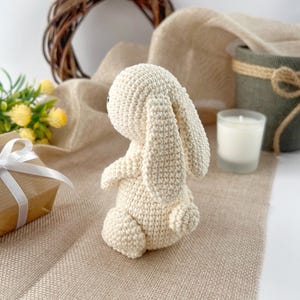 May include: A crocheted white bunny rabbit toy with long ears. The bunny is sitting on a light brown surface.
