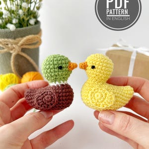 May include: Two crocheted ducklings, one brown and green, the other yellow, are held up. The green duckling has a white collar and orange beak. The yellow duckling has an orange beak. A circular graphic reads "PDF PATTERN IN ENGLISH".
