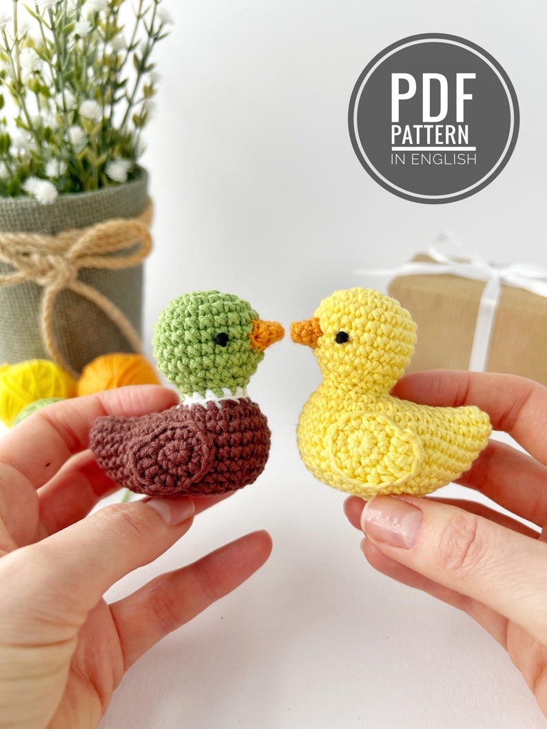 May include: Two crocheted ducklings, one brown and green, the other yellow, are held up. The green duckling has a white collar and orange beak. The yellow duckling has an orange beak. A circular graphic reads "PDF PATTERN IN ENGLISH".