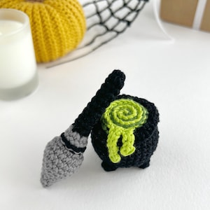 May include: A small black crocheted cauldron with a bright lime green swirled liquid bubbling over the rim, accompanied by a miniature grey and black crocheted broomstick. The items are crafted with visible yarn stitches and a textured finish.