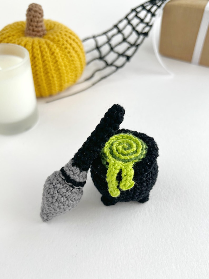 May include: A small black crocheted cauldron with a bright lime green swirled liquid bubbling over the rim, accompanied by a miniature grey and black crocheted broomstick. The items are crafted with visible yarn stitches and a textured finish.