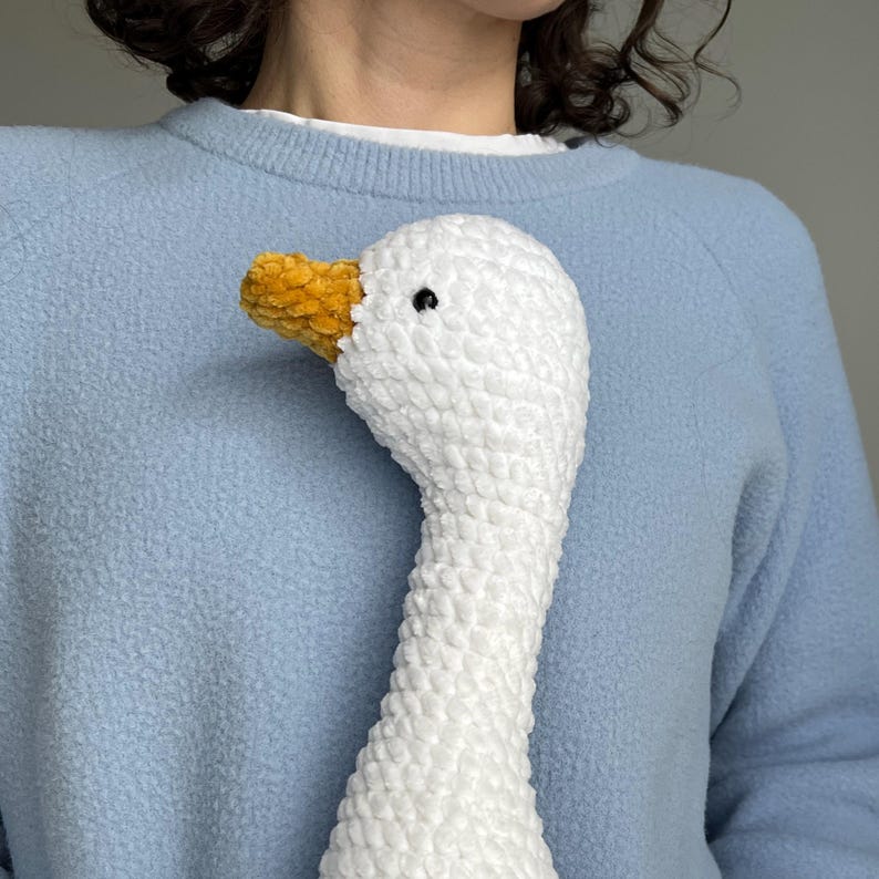May include: A white crochet goose with a yellow beak and black eye. The goose is made of a textured, plush material. The goose is against a light blue sweater.