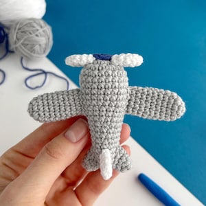 May include: A handmade gray crochet airplane with white propeller blades and tail. The airplane has blue accents and is held against a blue and white background. Yarn balls are visible in the background.