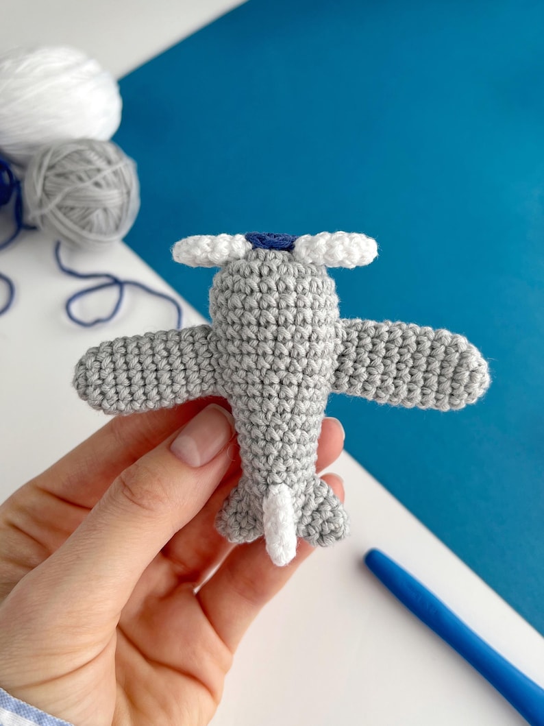 May include: A handmade gray crochet airplane with white propeller blades and tail. The airplane has blue accents and is held against a blue and white background. Yarn balls are visible in the background.