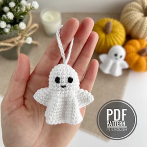 May include: A white crocheted ghost ornament with black eyes and a smiling mouth. The ghost has a loop for hanging. Other crocheted items in the background include pumpkins and a small ghost. The image includes the text "PDF PATTERN IN ENGLISH".