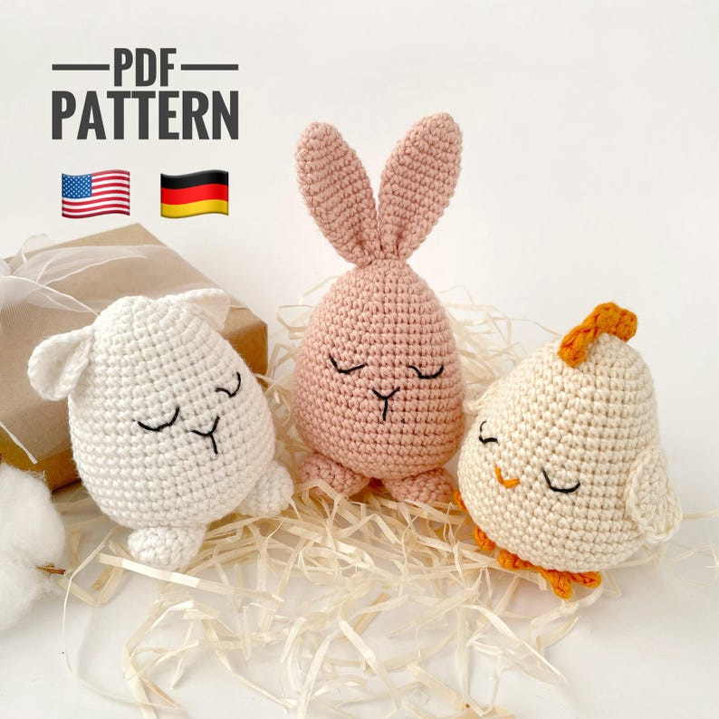 May include: Three crocheted stuffed animals: a pink bunny, a white lamb, and a white chick with an orange beak and feet. The animals have closed eyes and are sitting on a bed of straw. The text "PDF PATTERN" is above the animals, and the American and German flags are to the left of the text.
