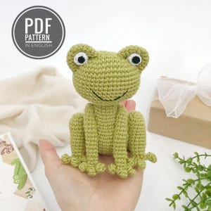 Può includere: Una rana verde all'uncinetto con occhi neri e bianchi e una bocca sorridente. La rana è seduta con le zampe distese. L'immagine include una grafica circolare con la scritta "PDF PATTERN IN ENGLISH".