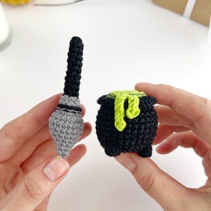 May include: A small crocheted black and grey broomstick alongside a crocheted black cauldron with a bright lime green liquid spilling over the rim. The cauldron features three small feet at the base and a textured, yarn-based construction.