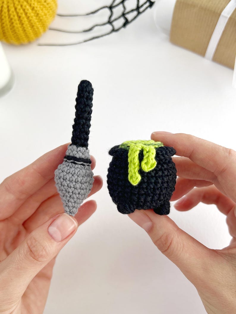May include: A small crocheted black and grey broomstick alongside a crocheted black cauldron with a bright lime green liquid spilling over the rim. The cauldron features three small feet at the base and a textured, yarn-based construction.