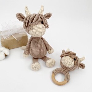 Puede incluir: Dos juguetes de vaca Highland de crochet en marr&oacute;n y blanco. Uno es un juguete de peluche m&aacute;s grande y el otro es un sonajero m&aacute;s peque&ntilde;o con un anillo de madera.