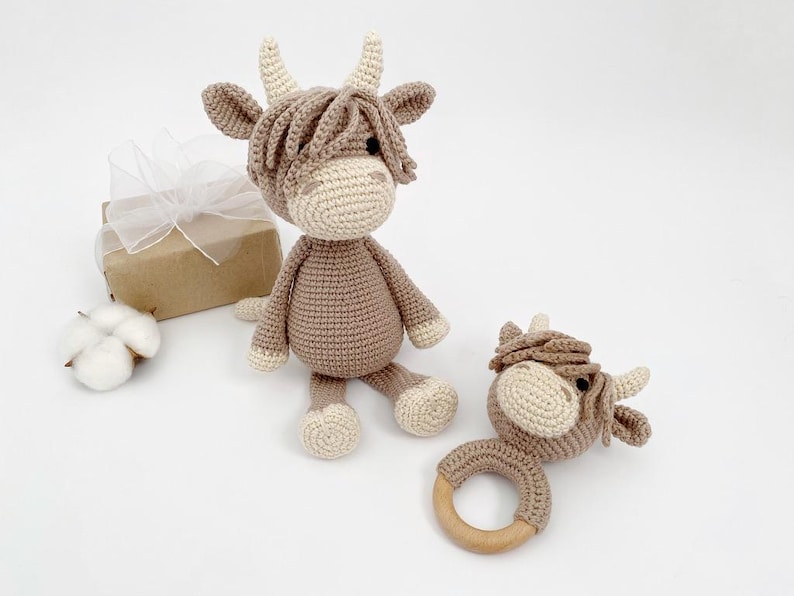 Puede incluir: Dos juguetes de vaca Highland de crochet en marr&oacute;n y blanco. Uno es un juguete de peluche m&aacute;s grande y el otro es un sonajero m&aacute;s peque&ntilde;o con un anillo de madera.