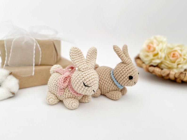 May include: Two crocheted bunny rabbits, one with a pink ribbon and the other with a blue ribbon. The bunnies are laying on a white surface.