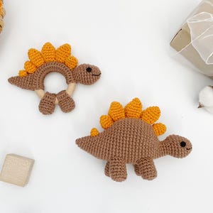 May include: Two crochet dinosaur toys in brown and orange. One is a rattle with a ring shape, and the other is a standing dinosaur. The toys have small black eyes and are on a white surface. A gift box and yarn are in the background.