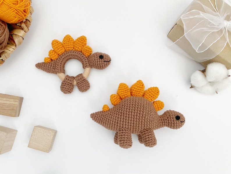 May include: Two crochet dinosaur toys in brown and orange. One is a rattle with a ring shape, and the other is a standing dinosaur. The toys have small black eyes and are on a white surface. A gift box and yarn are in the background.