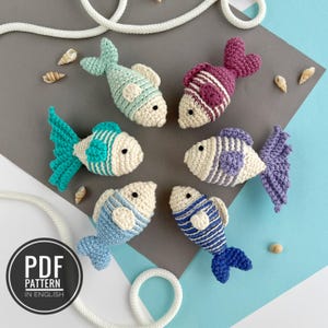 May include: Six colourful, handmade crochet fish in shades of blue, purple, and pink, with white detailing. The fish feature striped patterns and detailed fins. The image includes seashells and a rope, with a "PDF PATTERN IN ENGLISH" graphic.