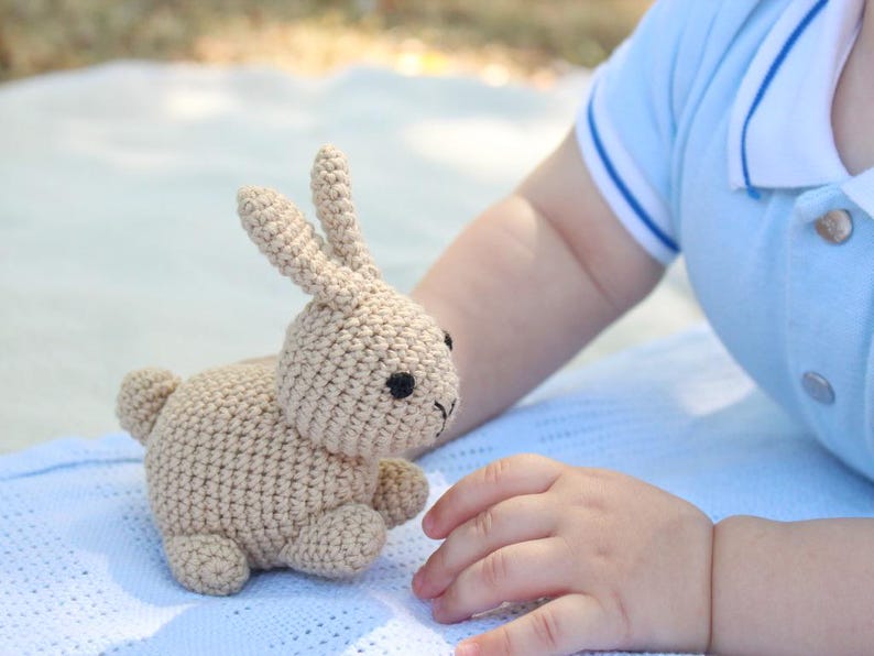 May include: A crocheted beige bunny toy with black eyes. The bunny is sitting on a blue blanket.