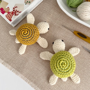 Puede incluir: Dos juguetes de tortuga hechos a mano con ganchillo. Cada tortuga tiene un cuerpo color crema y cuatro patas en forma de aleta. Una tortuga tiene un caparaz&oacute;n amarillo con un dise&ntilde;o en espiral, y la otra tiene un caparaz&oacute;n verde. Las tortugas est&aacute;n sobre una superficie beige.