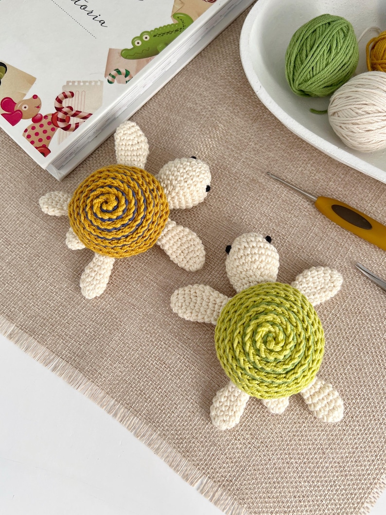 Puede incluir: Dos juguetes de tortuga hechos a mano con ganchillo. Cada tortuga tiene un cuerpo color crema y cuatro patas en forma de aleta. Una tortuga tiene un caparaz&oacute;n amarillo con un dise&ntilde;o en espiral, y la otra tiene un caparaz&oacute;n verde. Las tortugas est&aacute;n sobre una superficie beige.