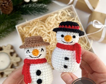 May include: Two small white crocheted snowmen figures. One snowman wears a brown hat and a red scarf, while the other wears a black hat with a red band and a red scarf. Both have orange noses, black eyes, stitched smiles, and two black buttons.