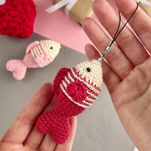 May include: Crocheted fish charm in red and cream with a black cord. The fish has a red tail and a cream-colored head. A second pink fish is in the background, along with a red heart and a wrapped gift.
