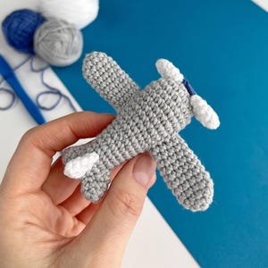 May include: A crocheted gray airplane with white accents on the wings and propeller. The handmade toy is held in a hand, with blue, white, and gray yarn balls in the background. The airplane is a charming, handcrafted item.